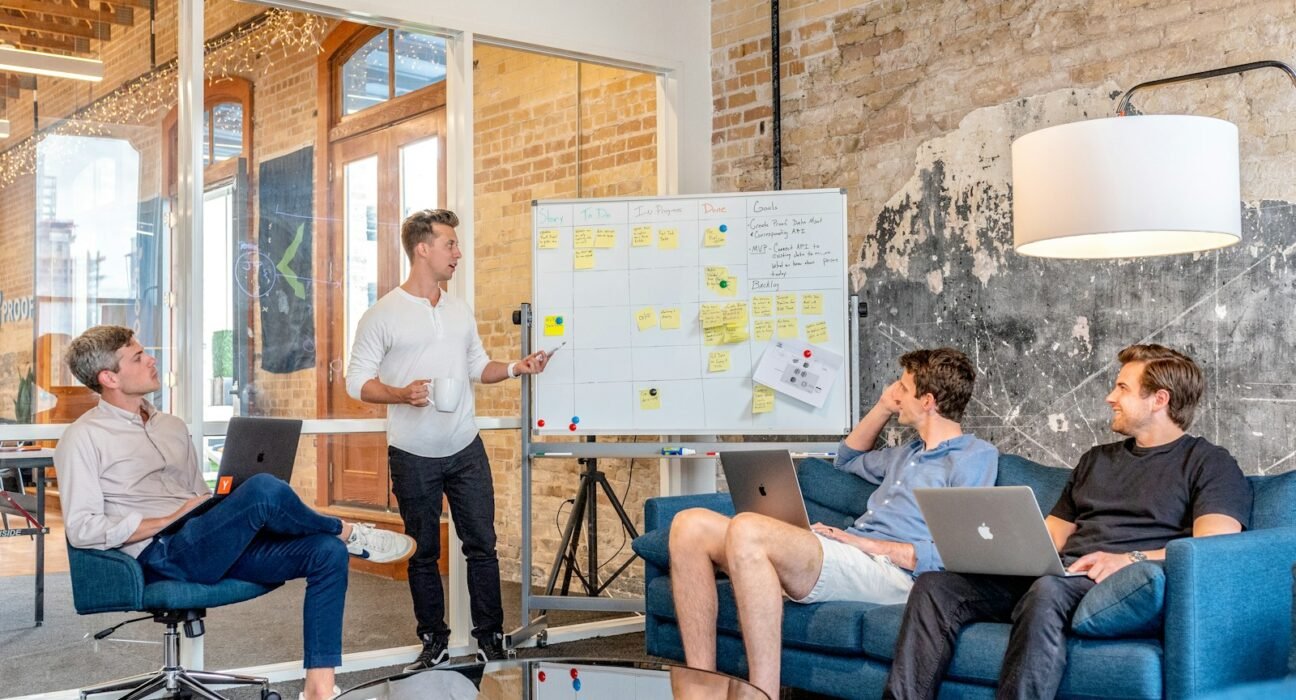 three men sitting while using laptops and watching man beside whiteboard