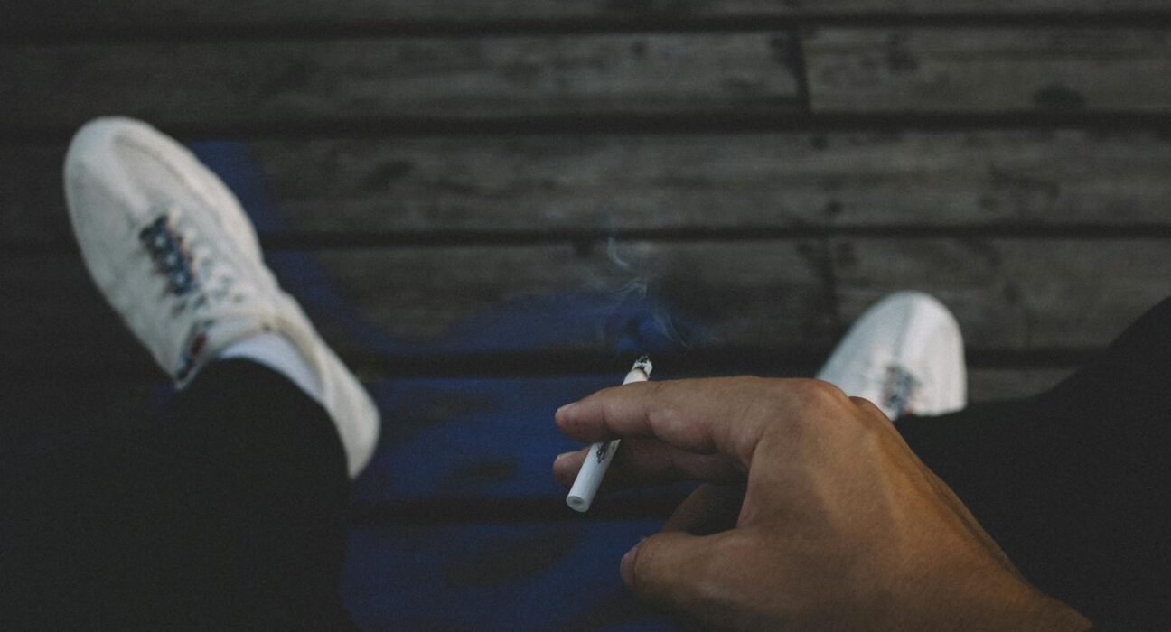 A man casually smoking a cigarette while relaxing outdoors on a wooden deck.
